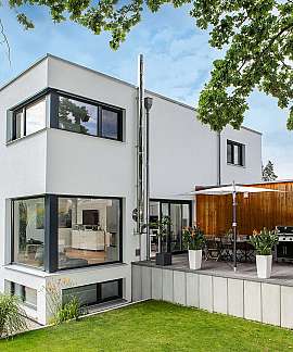 Modernes weißes Wohnhaus mit Holzterrasse, Garten und großen Fenstern unter Baumkronen.