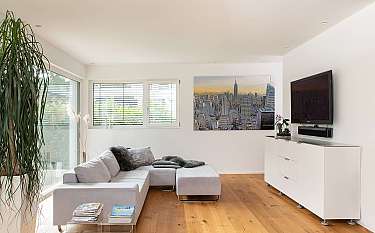 Helles Wohnzimmer mit grauem Ecksofa, Fernseher, weißem Sideboard und großem Fenster.