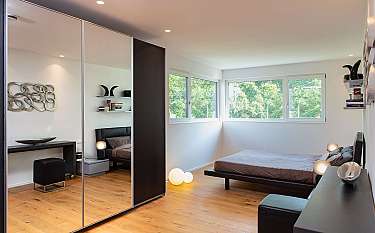 Helles modernes Schlafzimmer mit Holzfußboden, Spiegelkleiderschrank und Bett am Fenster.