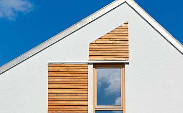 Weiße Hausfassade mit Holzpaneelen und einem Fenster vor blauem Himmel.