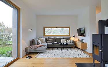 Helles Wohnzimmer mit grauem Ecksofa, großem Fenster, Teppich und schwarzem Kaminofen.