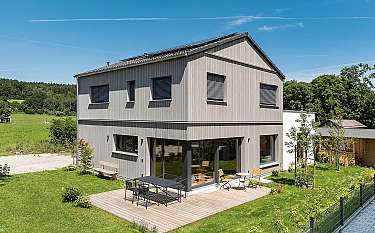 Zweigeschossiges modernes Haus mit Holzterrasse, Garten und Himmel im Hintergrund.