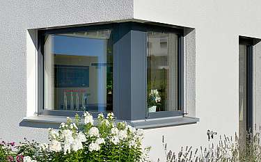 Großes Eckfenster an einer weißen Hausfassade mit Blumen im Vordergrund.