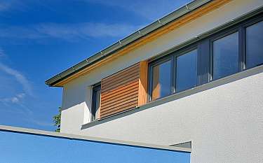 Weiße Hausfassade mit großen Fenstern, Holzverkleidung und blauem Himmel.