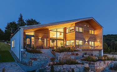 Modernes Holzhaus mit großen Fenstern und beleuchtetem Innenraum bei Abenddämmerung.