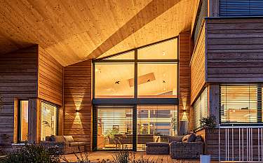 Holzverkleidete Hausfassade mit überdachter Terrasse, großen Fenstern und Sitzmöbeln im warmen Licht.