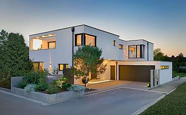 Moderne weiße Villa mit beleuchteten Fenstern, Garage und gepflegtem Vorgarten
