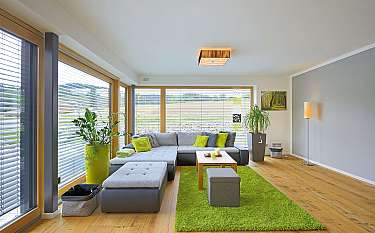 Helles Wohnzimmer mit Ecksofa, grünem Teppich, Pflanzen und großen Fenstern